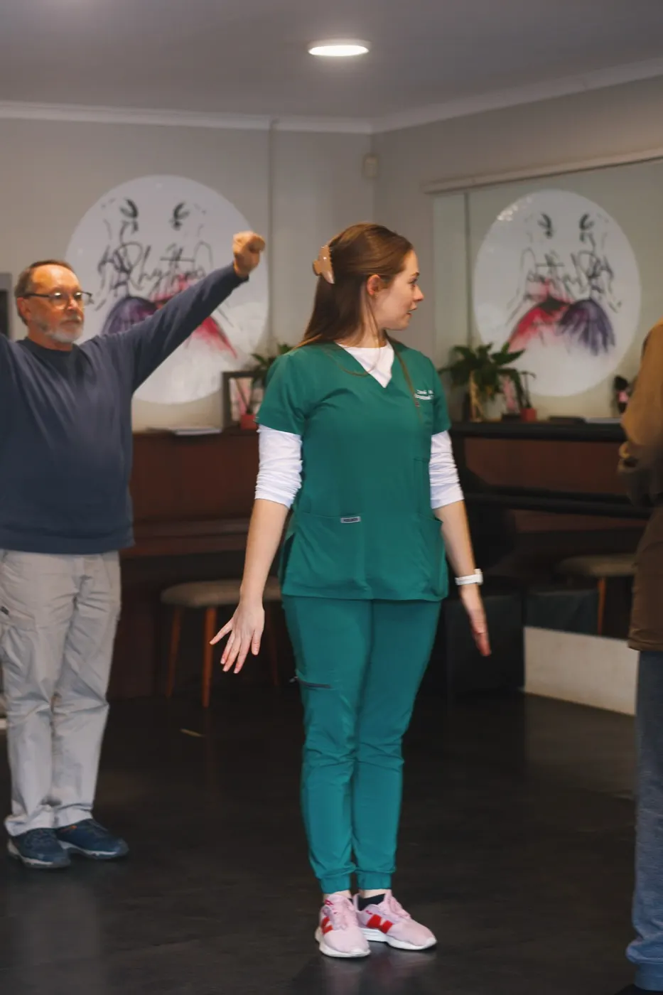 Therapist in green scrubs leading a standing arm raise while a participant follows behind.