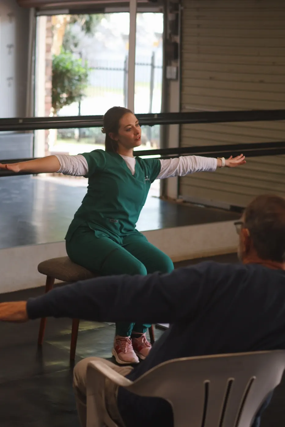 Therapist seated with arms stretched wide, demonstrating a movement to a participant.